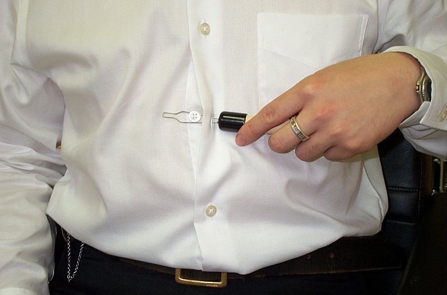 The diagram shows a person buttoning a shirt using button hook with only one hand.  The button has a handle and a metal loop at the tip.  When in use, first insert the metal hook into the button hole of the shirt, then use the metal hook to hook the button and finally pull the button through the button hole of the shirt.