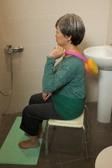 an elderly is sitting on a shower seat with a long handled brush. Her feet are placed on a non-slip mat