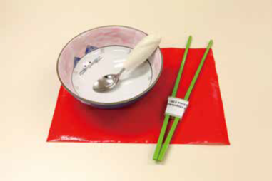 一张红色防滑垫, 上面放着一只碗，碗中有一只粗柄的匙, 旁边放着一对已套上筷子辅助器的筷子。